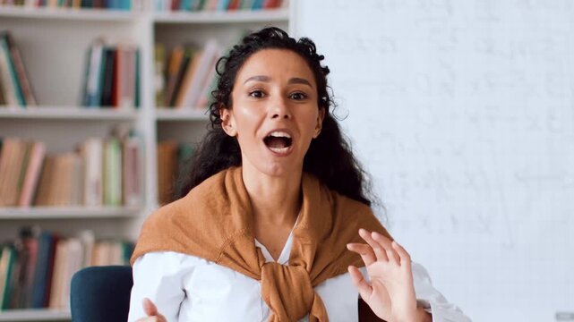 A woman with curly hair is sitting at a table with books in the background. She is smiling and appears to be discussing a topic with others.