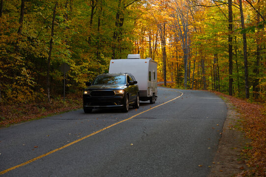 A black SUV towing a white travel trailer rounds a winding forested road through peak autumn foliage in Ontario, Canada, with headlights on. The surrounding hardwood trees blaze with orange, yellow,