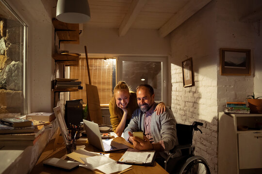 Smiling couple using smartphone and laptop at home office at night, man in wheelchair
