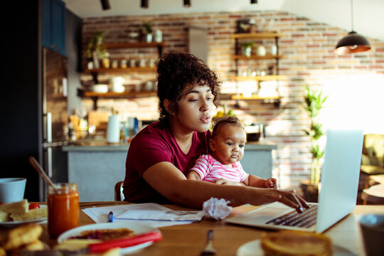 Mother working on laptop with baby at home kitchen table