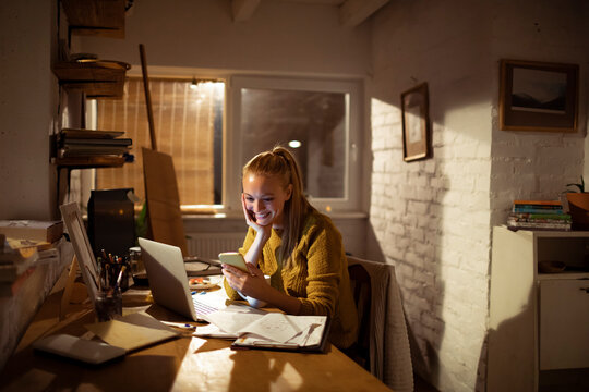 Smiling young woman using smartphone and laptop in home office at night