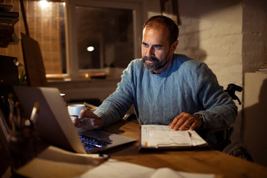 Middle-aged man in a wheelchair working on laptop at home office at night