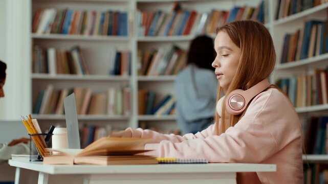 Two students are studying in a library. One student is wearing headphones and appears thoughtful while working on a laptop. The background shows shelves filled with books.