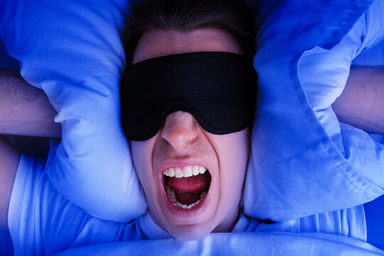 Angry man in a sleep mask screams while covering his ears with pillows in bed. Concept of insomnia, sleep deprivation, or loud noisy neighbors at night.