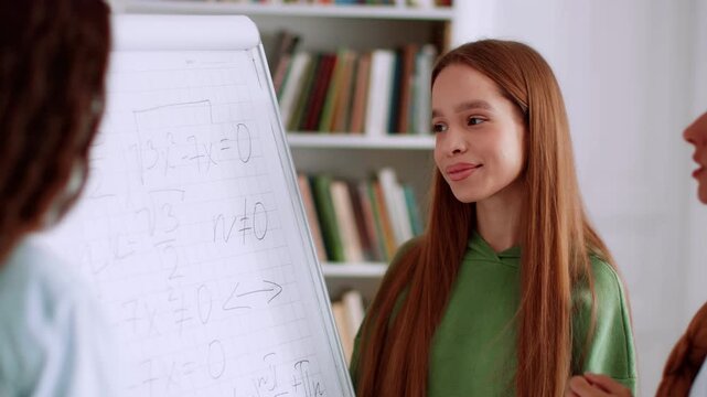 A young girl with long brown hair in a green hoodie stands next to a whiteboard. Two students share ideas while explaining math problems. Bookshelves are visible in the background.