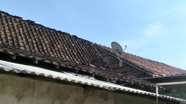 Weathered traditional clay tile roof on a residential building, featuring white satellite dish installed on the side under a clear blue sky. Condition of rural housing built with old building material
