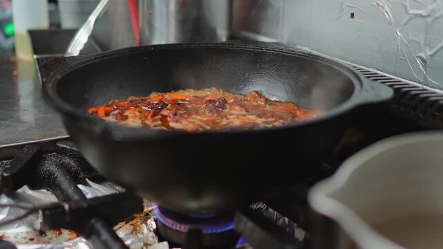 Side view of a black cast iron wok on a gas stove filled with simmering plov base of carrot, meat and spice with visible blue flame underneath, kitchen tools in background