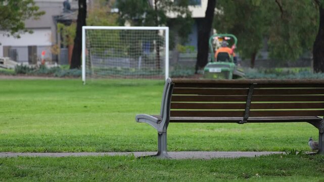 Park bench in foreground with a lawn mowing worker maintaining grass near a soccer goal in background in Melbourne Australia. Public park showing maintenance activity, community recreational space.
