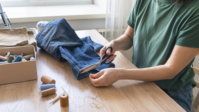 Hands cutting thread from old jeans for reuse, promoting sustainable fashion and circular economy principles. Person upcycling denim jeans with scissors, repairing clothing