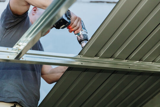 A mature roofer installs roofing sheets on a profile shed using a drill.