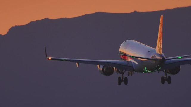 Close up side view of commercial airliner with red orange tail fin on final approach, landing gear down, vivid orange sunset sky and mountain range silhouette in background, green navigation light on