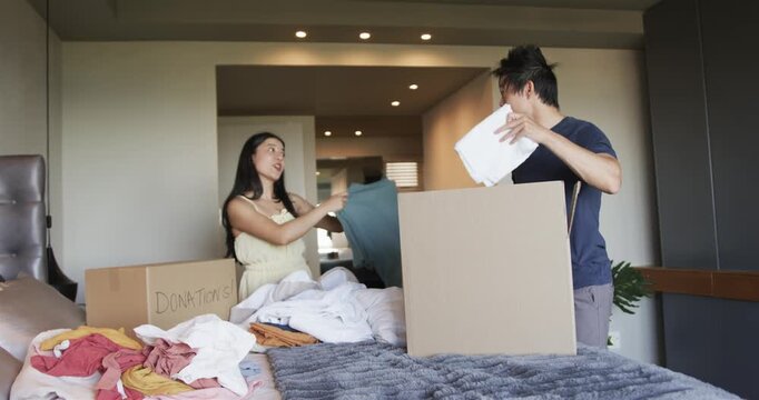 Asian couple sorting and packing clothing in bedroom by bed with donations box and teal top