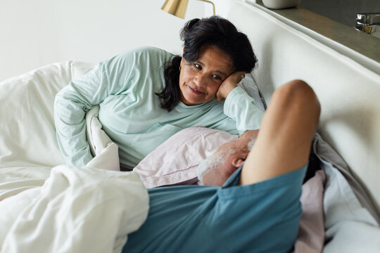 Senior African American couple reclining in bedroom on bed with white duvet, woman gazing at man