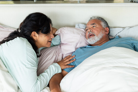 Senior couple lying in bed in bedroom, woman leaning over man, touching chest, wearing mint top