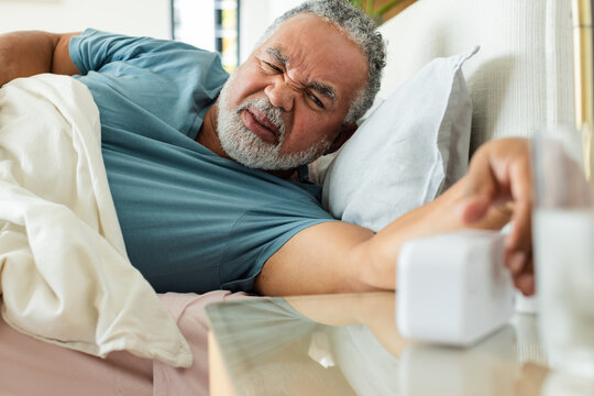 Senior African American man in teal tee reaching clock while ringing, getting water, waking in bed