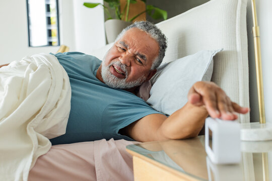 Senior African American man lying in bed wearing blue shirt reaching for alarm clock on nightstand