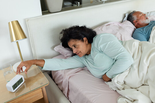 Senior African American couple lying in bedroom, woman in green pajama top reaching alarm clock