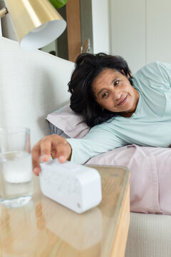 Mature African woman reclining in bedroom wearing light blue top reaching toward white alarm clock