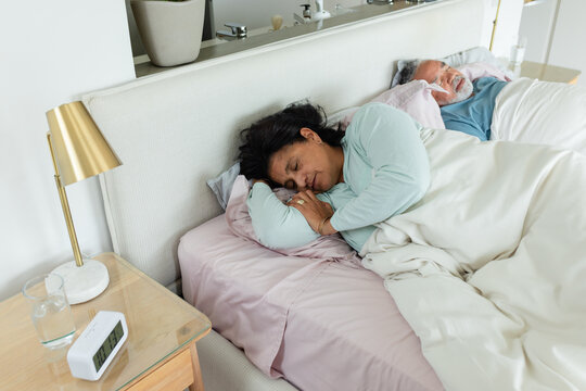 Mature African American couple in bedroom with woman sleeping on left under duvet as alarm buzzing