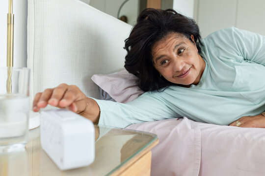Mature African woman lying on pale pink bedding, reaching for white device on glass-top nightstand