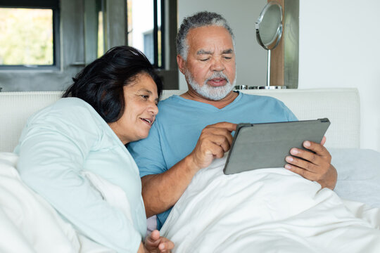 Senior African American couple lying under white duvet on bed, man holding tablet in folio resting