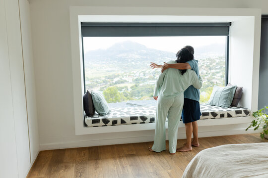 African American couple standing at picture window in bedroom pajamas pointing at hills