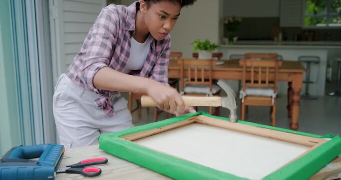 African woman finding frame hammering to secure green fabric, inspecting panel upright on worktable