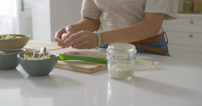 Asian woman peeling hard-boiled egg on countertop while prepping salad with sprouts and scallion