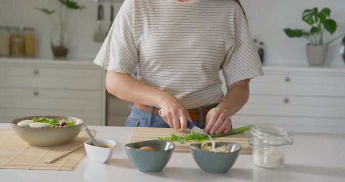 Woman placing, slicing scallion with chef knife on board at kitchen, striped shirt prepping salad