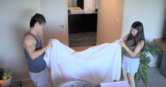 Asian couple, man lifting sheet from basket, shaking, smoothing, folding by bed to join towel stack