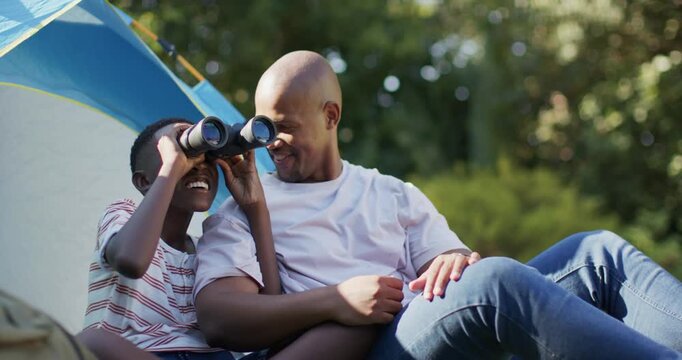 African father and teenage son lifting binoculars, guiding, scanning for birds at camp by blue tent