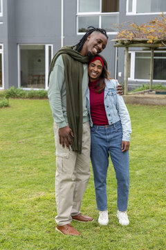 Indian and African American couple standing on grassy lawn by gray building, wearing denim jacket