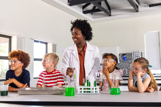 Adult female educator in lab coat leading chemistry demo at lab bench, four children in goggles