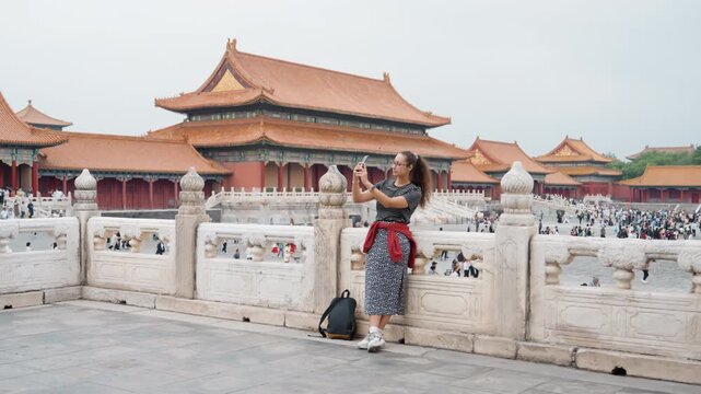 Young woman posing at forbidden city stone balustrade overlooking palace rooftops, patterned dress and backpack, quiet travel moment with distant crowd, soft overcast light highlighting ornate eaves