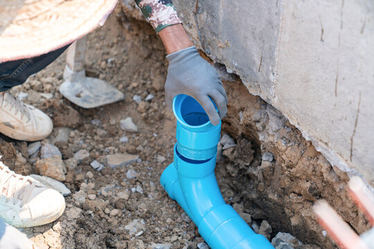 Plumber installing blue PVC drainage pipe system in a ground trench for building construction.