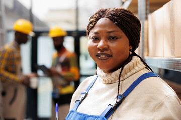 Fototapeta premium Smiling african american woman working in industrial warehouse portrait. Shipment manager with positive facial expression standing near parcels shelf and looking at camera close up
