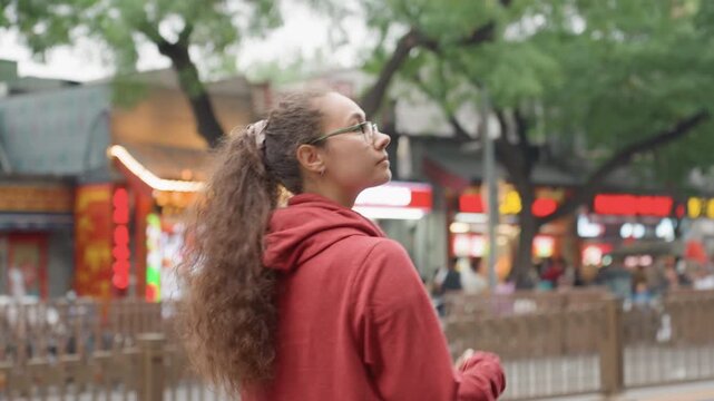Young woman red hoodie exploring city market, backpack over shoulder, curly ponytail and glasses, walking along sidewalk past storefronts and trees, soft bokeh lights and distant neon, candid profile