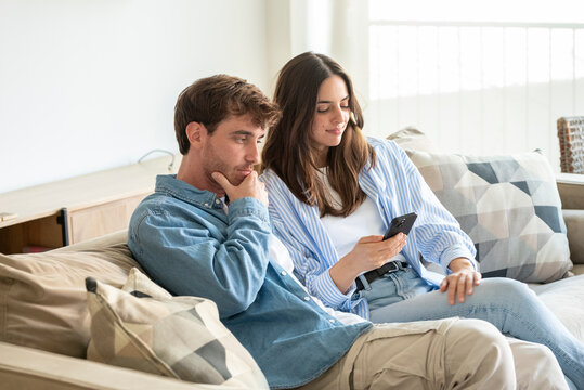 A young couple sits on a bright living room sofa, sharing a smartphone and smiling as they browse content. Casual clothing, cozy cushions and everyday intimacy capture a modern relationship moment.