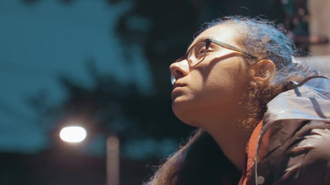Young woman with snack looks upward. Portrait of woman holding snack under lamplight in city night. Nighttime city street capturing young woman gazing upward with contemplative expression