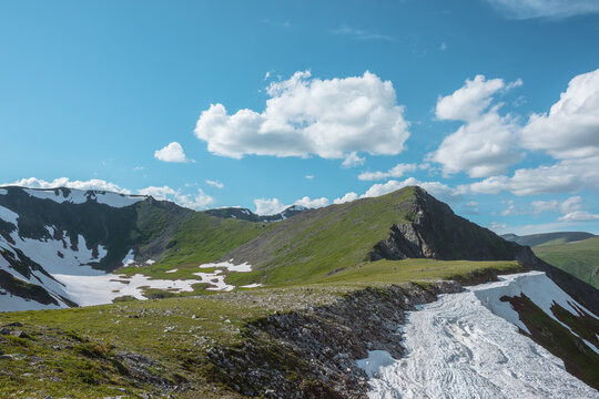Scenic sunlit view along grassy ridge with white snow cornice to green hill top with rocky sheer crags in bright sun under lush clouds in blue sky. Vivid alpine scenery in high mountains in sunny day.