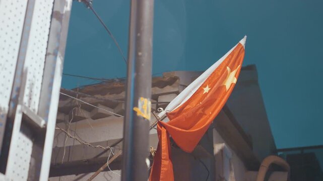 Sunlit red china flag fluttering rooftop, closeup showing worn fabric snagged on metal pole, weathered eaves and tangled utility wires, clear blue sky backdrop, vintage film grain and warm toning