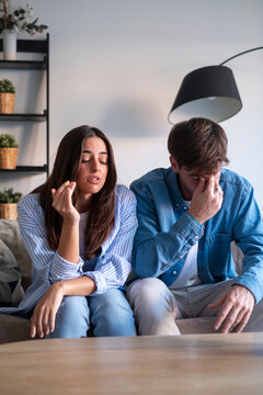 A young couple sits on a couch after an emotional argument, both appearing stressed and frustrated as they struggle with relationship conflict and emotional exhaustion in a modern living room.