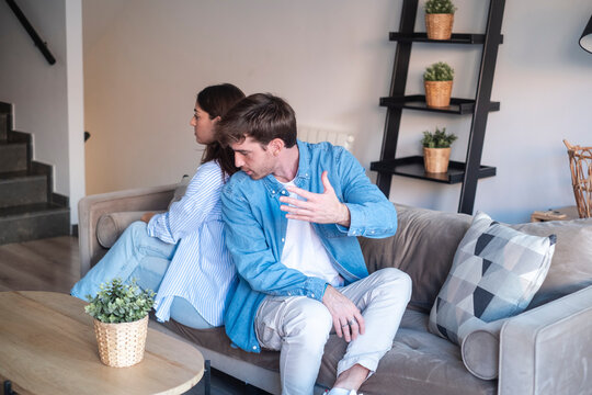 A young couple sits back-to-back on a sofa after an argument, showing tension and emotional distance in a contemporary living room setting with neutral decor and casual clothing.