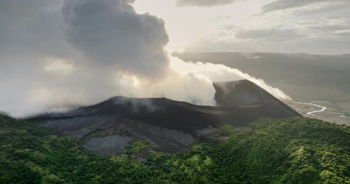 Mount Yasur volcano on Tanna Island erupting, thick white smoke and ash pouring from crater over dark volcanic slopes, framed by lush green vegetation and a river below. Aerial zoom out drone panorama