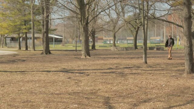 Man walking toward camera through Kew Gardens park among bare trees in early spring, Toronto, Canada.