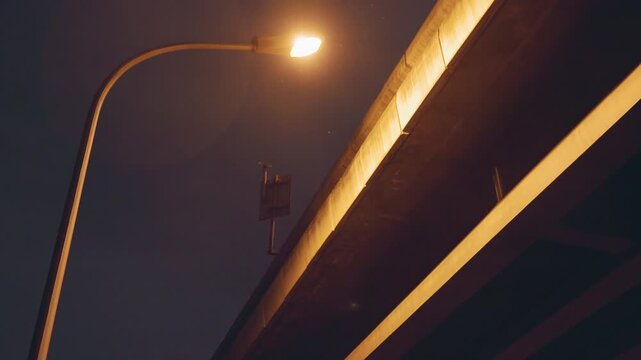 Night overpass illuminated by lamp, golden glow on concrete underside of bridge, empty urban scene with distant surveillance fixture, cinematic shadow and quiet industrial atmosphere