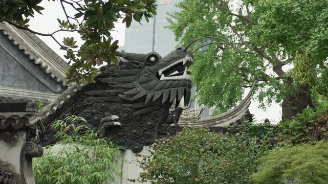 Ornate dragon roof carving peeking through garden foliage, black ceramic scales and open maw with dripping detail urban skyline glimpsed beyond, moody light and protective aura suited for cultural