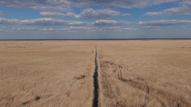 A wide aerial view of an endless dry grassland with a canal stretching straight toward the horizon. The expansive sky filled with soft clouds enhances the sense of openness and solitude.