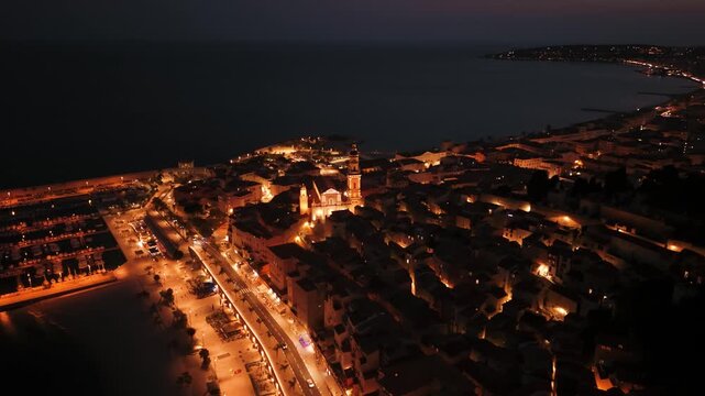 Menton Basilique Saint-Michel glowing over old town with harbor and coastal jetty at night, France, aerial
