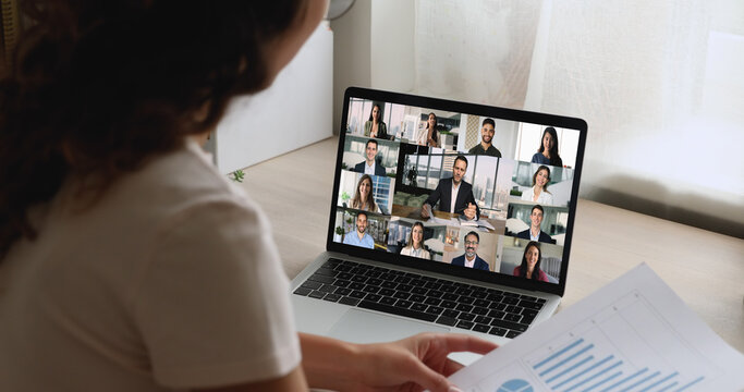 Businesswoman engages in virtual meeting with diverse group displayed on laptop screen, view over her shoulder. Integration of technology for professional communication in globally connected workspace
